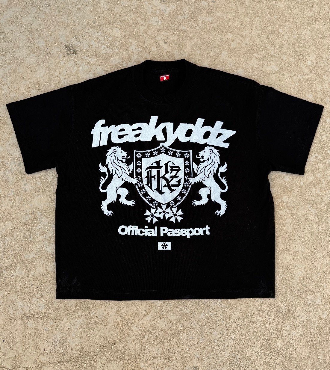 Camiseta FREAKYDDZ Official Passport, escudo con dos leones y lema Official Passport, sobre fondo de cemento